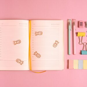 Flat lay of colorful office supplies with a notebook on a pink background. Ideal for workspace inspiration.