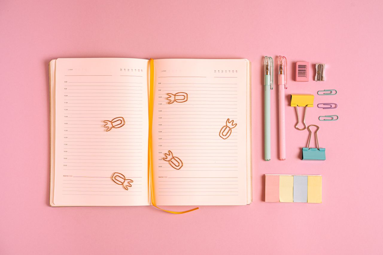 Flat lay of colorful office supplies with a notebook on a pink background. Ideal for workspace inspiration.